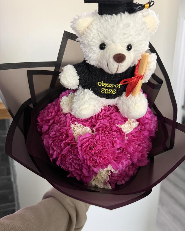 Grad Bear Ramo by Blooms x Mel — Hot pink carnations topped with a Class of 2026 bear holding a diploma, in a black wrap