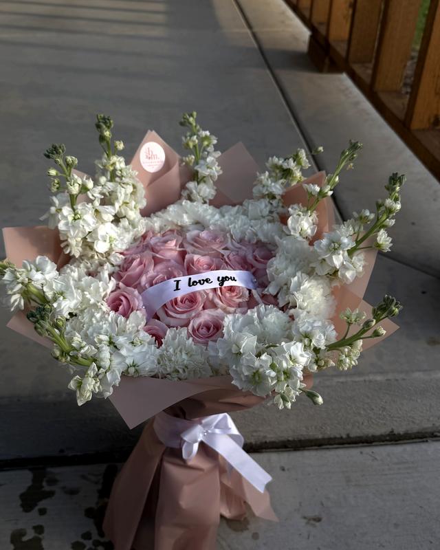 The I Love You by Blooms x Mel — Pink roses circled in white carnations and stock with an "I love you" banner across the center